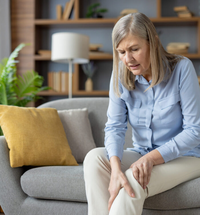 Pensioner woman sitting on grey couch and holding knee while bending over with grimace of pain. Suffering lady holding leg with inflammation related to ageing process and joint degeneration.