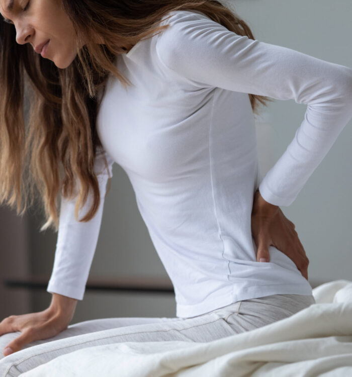 Close up unhappy woman wearing pajama rubbing stiff back muscles after awakening, sitting on bed, upset young female feeling pain, incorrect posture or uncomfortable bed, backache after sleep