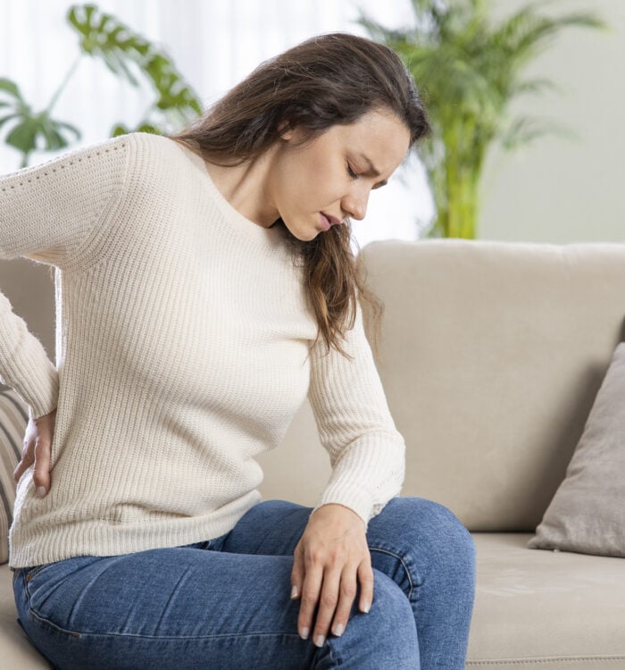 Young woman suffering with back pain, sitting on a couch and holding her lower back with hand. Axial pain, backache